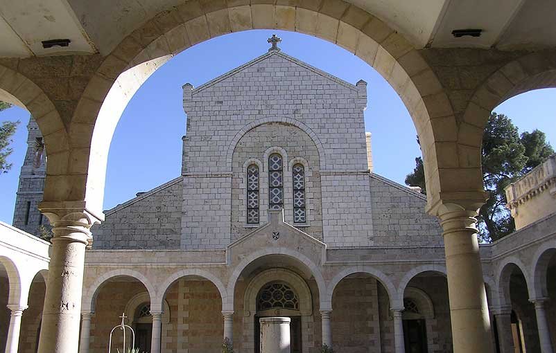 L'image montre un cloître avec des arches stylisées entourant un jardin. Au centre, on peut voir une structure architecturale en pierre, probablement une église ou un monastère, avec un toit en pente et des fenêtres ornées. L'ambiance est paisible, avec une lumière naturelle qui illumine le décor, créant un contraste entre l'ombre des arches et la clarté extérieure. Des éléments de verdure, comme des plantes, ajoutent une touche de vie à l'environnement.