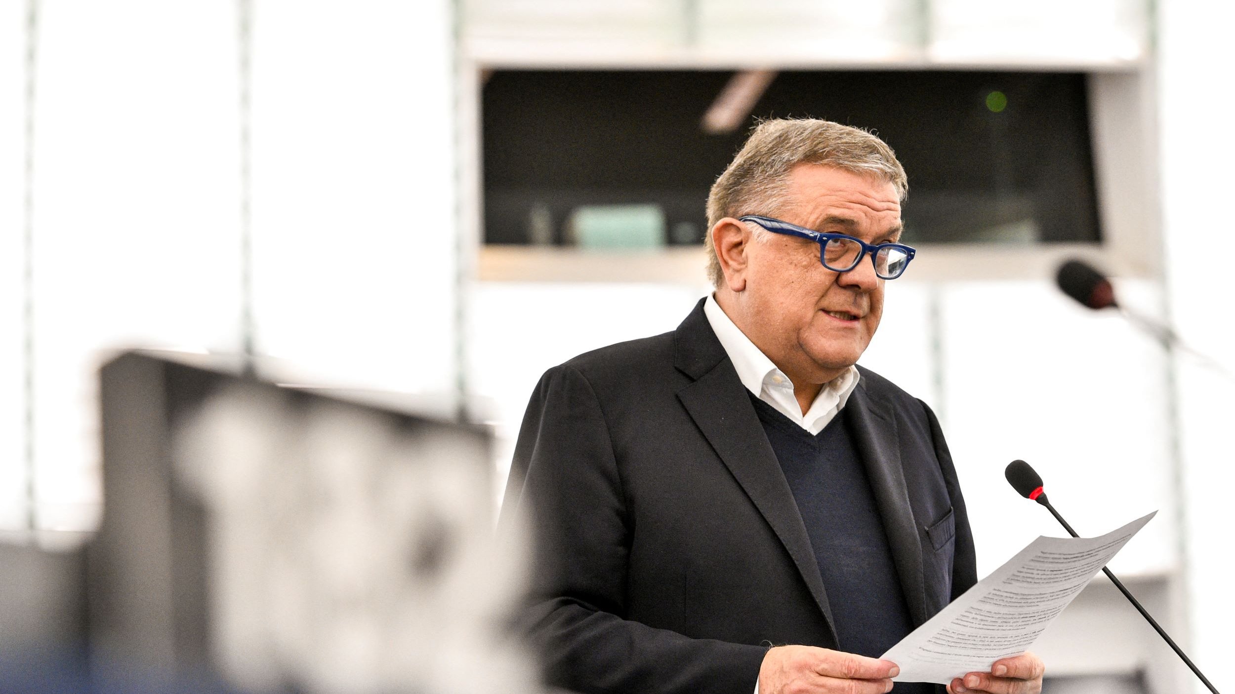 L'image montre un homme qui parle au micro lors d'une session au Parlement. Il porte des lunettes bleues et un costume sombre. Il semble lire un discours ou un document. L'environnement est celui d'une salle de réunion institutionnelle, avec un éclairage moderne et des panneaux en arrière-plan.