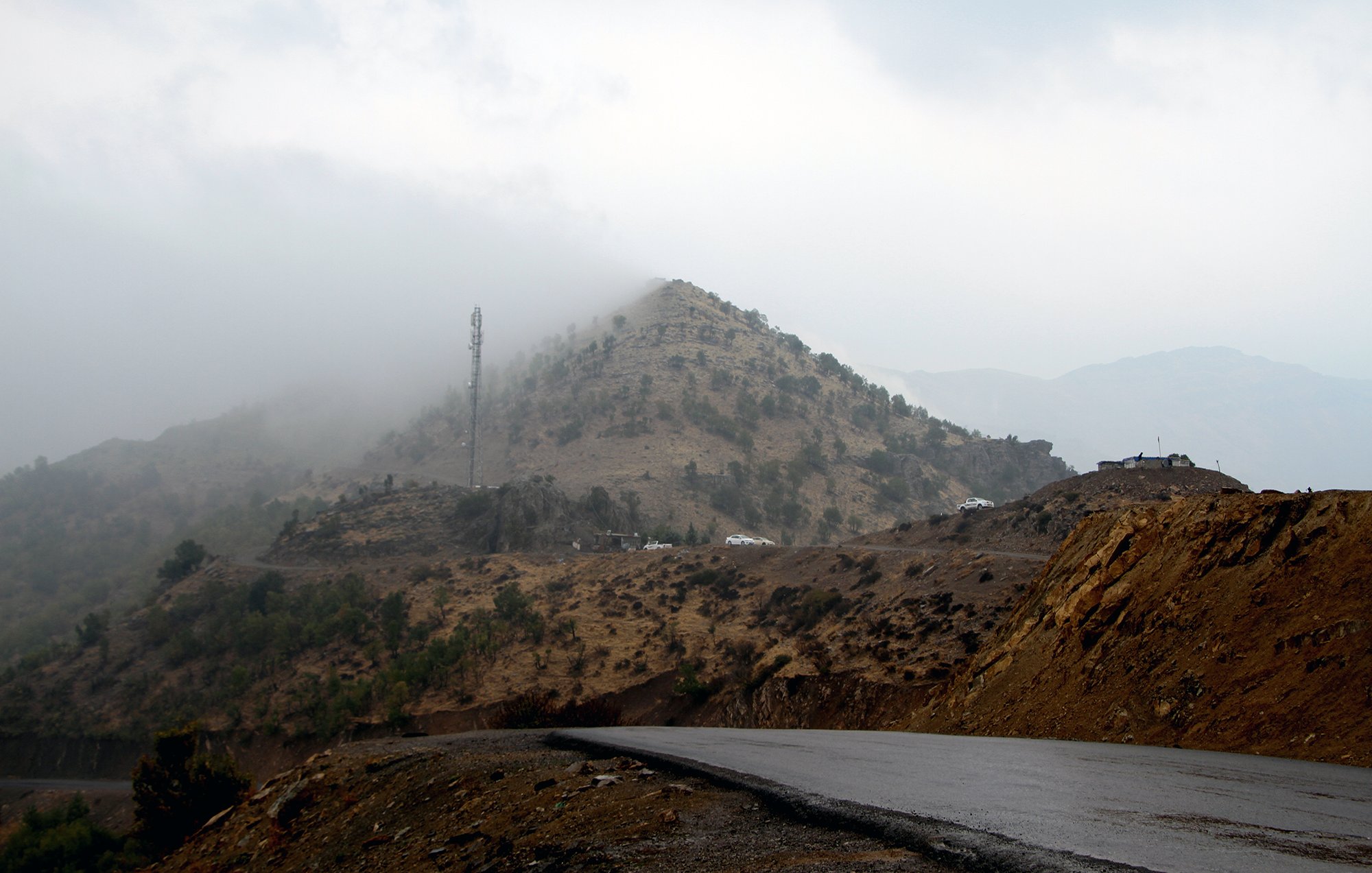 The image depicts a winding road leading through a mountainous landscape. In the background, a hill covered with low vegetation is partially obscured by mist or fog, creating a mysterious atmosphere. Atop the hill, there is a communication tower, and some vehicles are visible near the summit. The foreground features a dark, asphalt road that curves, suggesting a remote and rugged terrain. The overall scene conveys a sense of tranquility and solitude in nature.