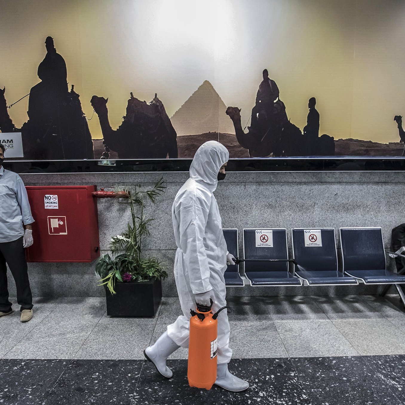 The image depicts a scene in a public area, likely an airport or a similar facility. In the foreground, a person wearing a white protective suit and mask is walking while holding an orange container. In the background, a large advertisement features silhouettes of camels and a pyramid, suggesting a connection to Egypt. There are two individuals in the background, also wearing masks, standing near a fire extinguisher. The seating area appears empty, adding to a sense of quietness in the environment.