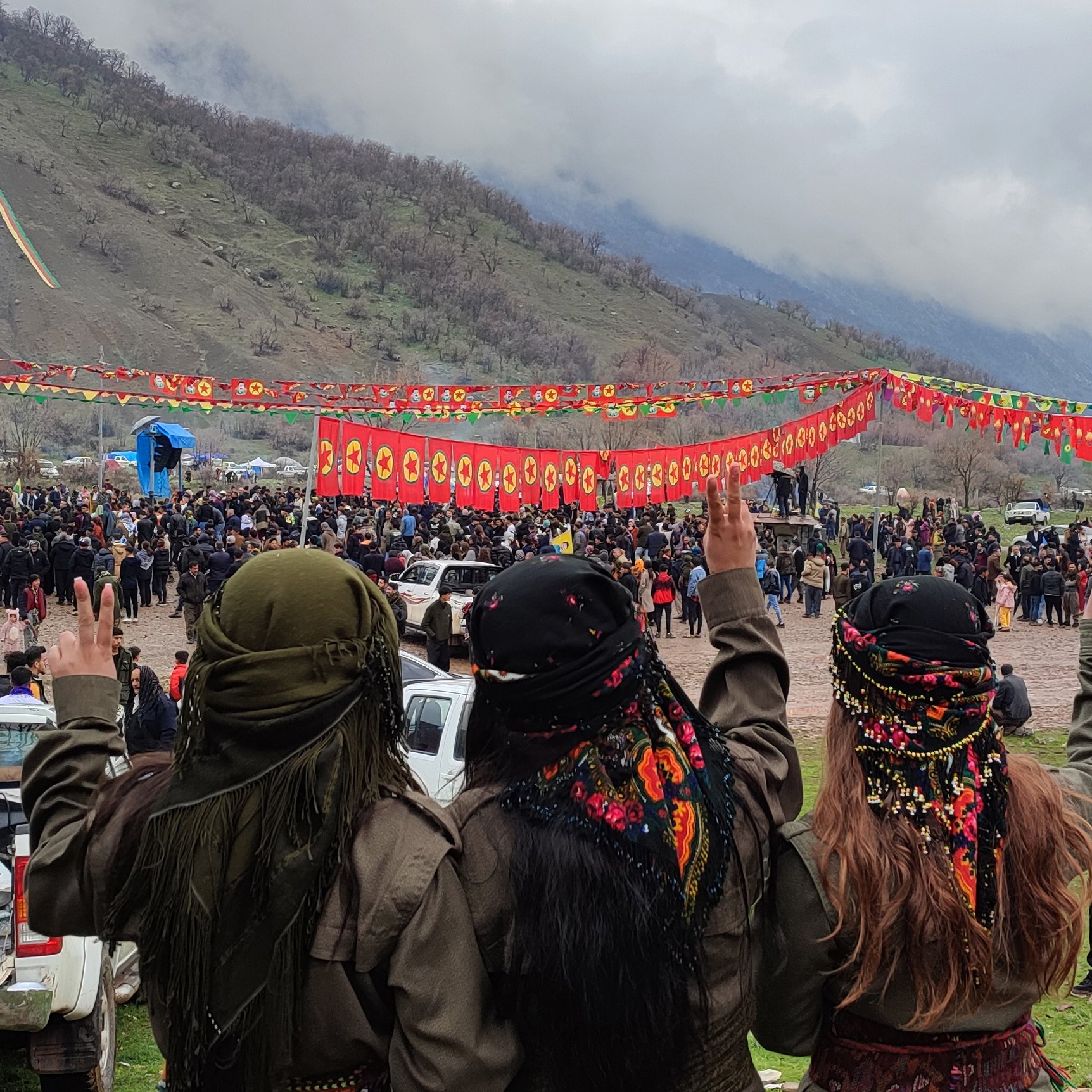 L'image montre un groupe de trois personnes vues de dos, faisant un signe de paix avec leurs mains. Elles portent des vêtements traditionnels et des foulards colorés. En arrière-plan, on peut voir une foule rassemblée pour un événement, avec des banderoles rouges décorées de symboles. Le paysage environnant est montagnard et sous un ciel nuageux. L'atmosphère semble festive et solennelle.