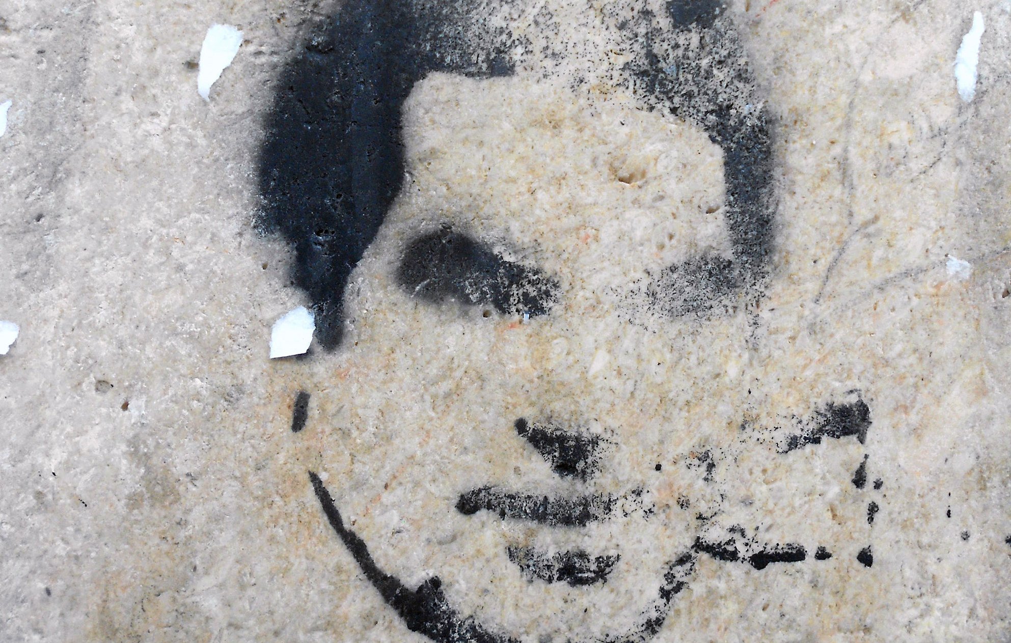 L'image montre un pochoir sur un mur, représentant un visage stylisé. Les contours sont en noir, avec un fond de pierre claire. Le visage semble être celui d'une personne jeune, avec une expression neutre. Quelques résidus de papier sont éparpillés autour, ajoutant une texture supplémentaire au mur. L'ensemble dégage une atmosphère urbaine et artistique.