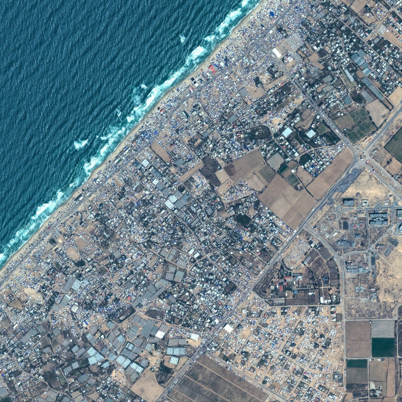 L'immagine mostra una vista aerea di un'area costiera. Si possono osservare le onde del mare che si infrangono sulla riva, evidenziando una linea di costa ben definita. L'area terrestre è caratterizzata da un mix di edifici e infrastrutture, con una disposizione varia di strade e terreni agricoli. I colori variano tra il blu dell'acqua, il verde delle aree coltivate e il grigio dei centri abitati e delle strutture urbane. L'immagine presenta un contrasto interessante tra le zone urbanizzate e quelle naturali.