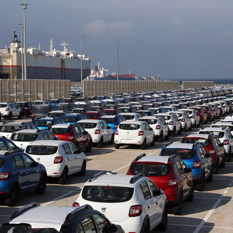 L'image montre un grand parking de voitures, organisé en rangées bien alignées. On aperçoit plusieurs types de voitures de différentes couleurs, majoritairement en blanc, rouge et bleu. En arrière-plan, on peut voir des bateaux de marchandises amarrés dans un port, sous un ciel nuageux. Cette scène semble se dérouler dans une zone portuaire, où les véhicules sont probablement en attente d'expédition ou de distribution.