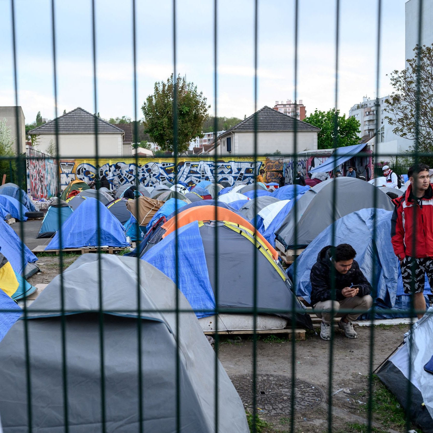 L'image montre un campement de tentes installé dans une zone urbaine. Les tentes sont de différentes tailles et couleurs, principalement en bleu et gris. On peut voir des personnes présentes, certaines debout et d'autres assises, semblant utiliser leur téléphone. En arrière-plan, il y a des bâtiments et un mur décoré de graffitis. L'ensemble du camp est entouré par une clôture, ce qui donne un aspect de confinement à la scène. Les arbres et la végétation ajoutent une touche de verdure au décor.