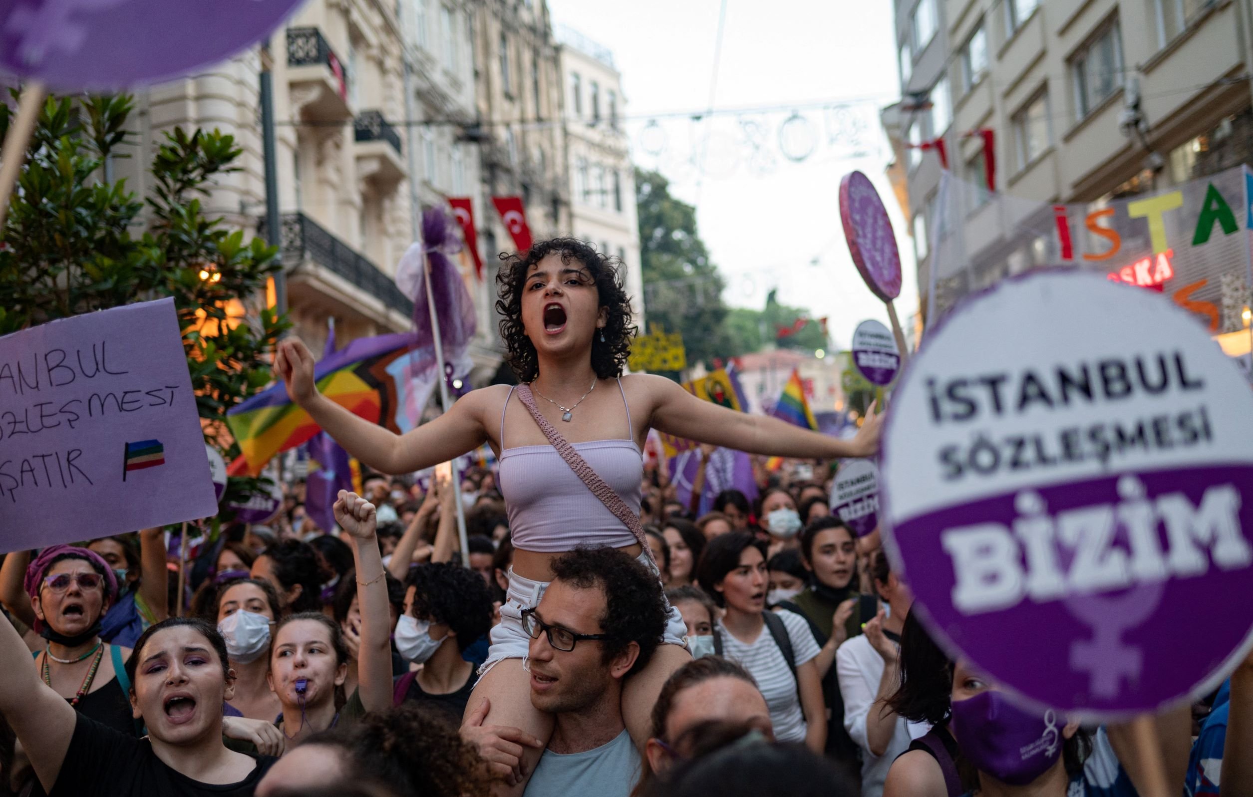 L'immagine mostra un gruppo di persone in una manifestazione per i diritti, con un forte focus sulle tematiche femministe e LGBTQ+. Una giovane donna, in primo piano e con i capelli ricci, è in piedi sulla spalle di un'altra persona e grida, esprimendo la sua passione. Molti partecipanti, prevalentemente donne, sono circondati da bandiere e cartelli colorati, alcuni con slogan che richiamano l'attenzione su temi di uguaglianza e diritti umani. L'atmosfera è vibrante e carica di energia, con un chiaro senso di solidarietà tra i manifestanti.