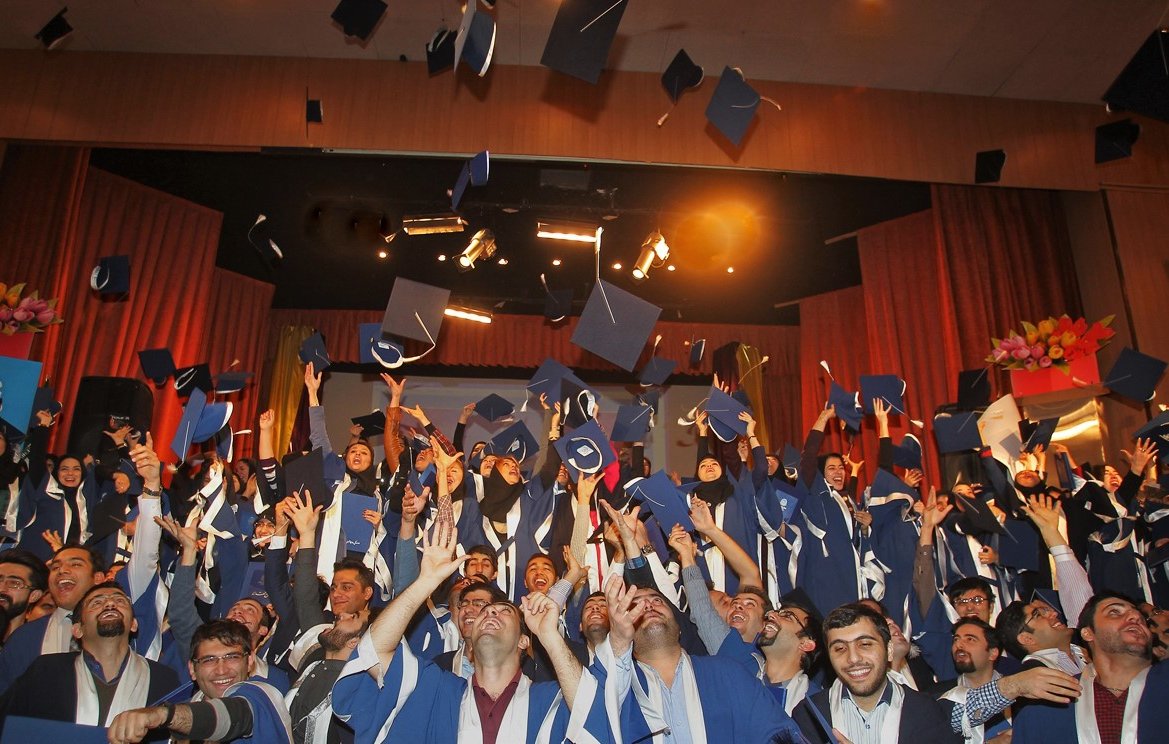 L'image montre une célébration de remise de diplômes. De nombreux étudiants, vêtus de toges et de mortiers de graduation, se tiennent ensemble sur scène. Ils lèvent leurs chapeaux en l'air, exprimant leur joie et leur excitation. L'atmosphère est festive, avec des sourires sur les visages des diplômés, soulignant un moment marquant de leur parcours académique. Des lumières et des décorations de scène ajoutent à l'ambiance festive.