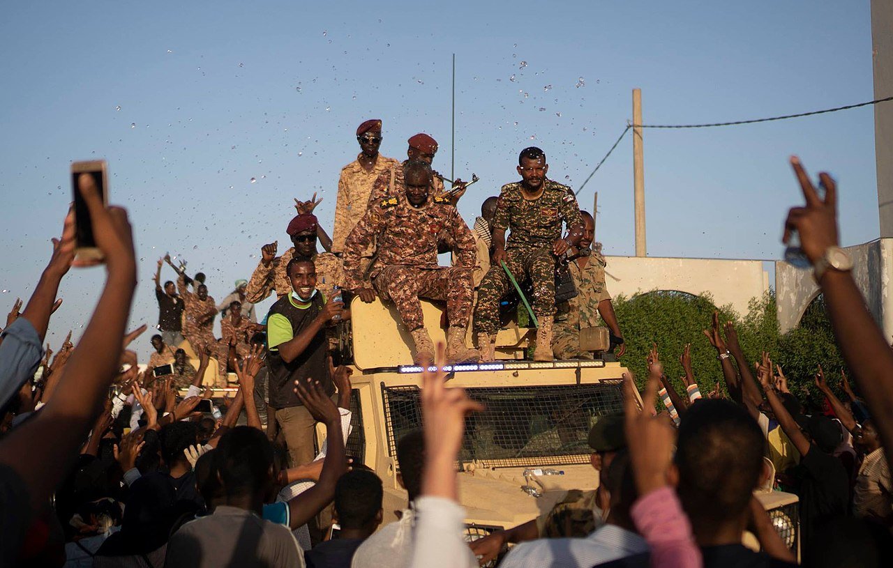 L'image montre un groupe de personnes en liesse rassemblées autour d'un véhicule militaire. Des soldats, vêtus d'uniformes camouflés, sont visibles en hauteur sur le véhicule, tandis que la foule en dessous lève les bras et semble manifester de la joie ou de l'enthousiasme. On peut apercevoir des confettis ou des éclats de couleur qui ajoutent à l'ambiance festive. L'atmosphère générale semble être celle d'une célébration ou d'un événement important.