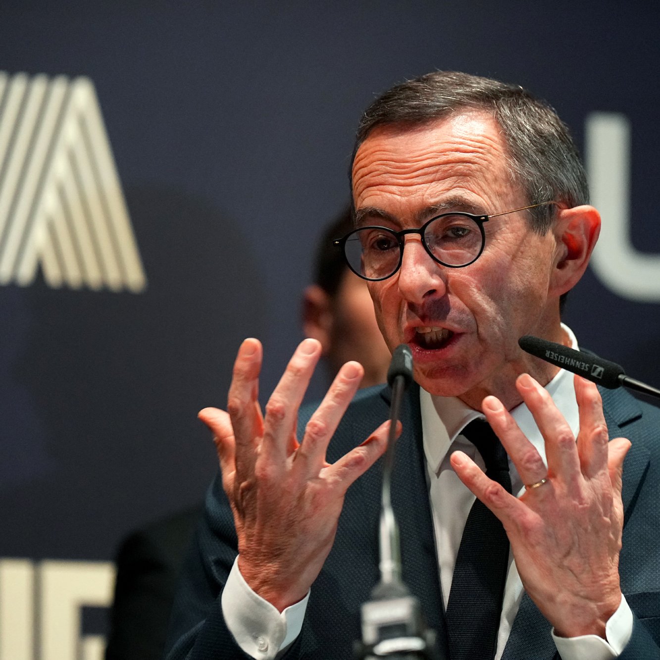 L'image montre un homme en train de parler lors d'un événement. Il porte des lunettes et a une expression animée, utilisant probablement des gestes pour renforcer son discours. En arrière-plan, on peut voir un logo qui semble être lié à une organisation, et des silhouettes floues de personnes apparaissent derrière lui. L'ambiance semble être professionnelle, suggérant que cet homme est en train de s'adresser à un public ou à des médias.