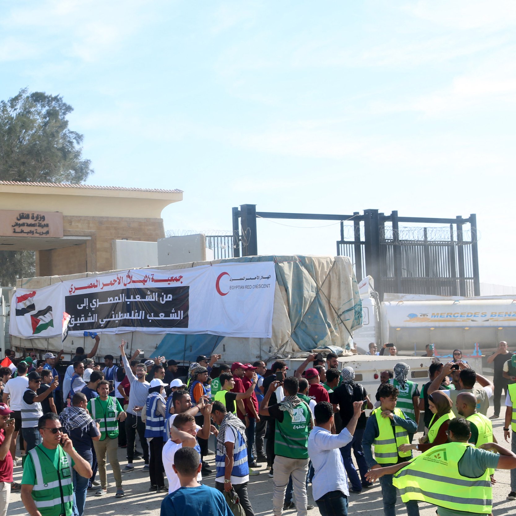 در تصویر، تعدادی از افراد در حال برگزاری یک تجمع یا اعتراض هستند. مردم در جلو ورودی یک ساختمان بزرگ ایستاده‌اند و برخی از آنها جلیقه‌های شرایطی به تن دارند. در پس‌زمینه، بنری بزرگ با متنی به زبان عربی و تصاویری از علم‌ها به چشم می‌خورد. همچنین در بالای این ساختمان، علم‌های کشورها در اهتزاز هستند. فضای تجمع به نظر شلوغ و پر انرژی می‌رسد.