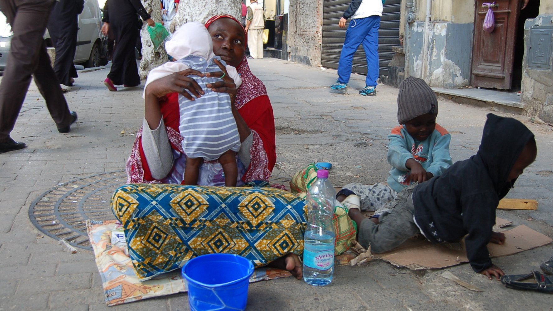 L'image montre une femme assise sur le sol, tenant un bébé dans ses bras. Elle est entourée de deux enfants, l'un assis à côté d'elle et l'autre jouant sur le sol. La femme porte un vêtement coloré et un foulard, tandis que les enfants semblent habillés de manière simple et modeste. À côté d'eux, il y a une bouteille d'eau et un seau bleu. La scène se déroule dans une rue, avec des bâtiments en arrière-plan et d'autres personnes qui passent. L'ambiance semble à la fois familiale et difficile, évoquant des thèmes de pauvreté et de lutte quotidienne.