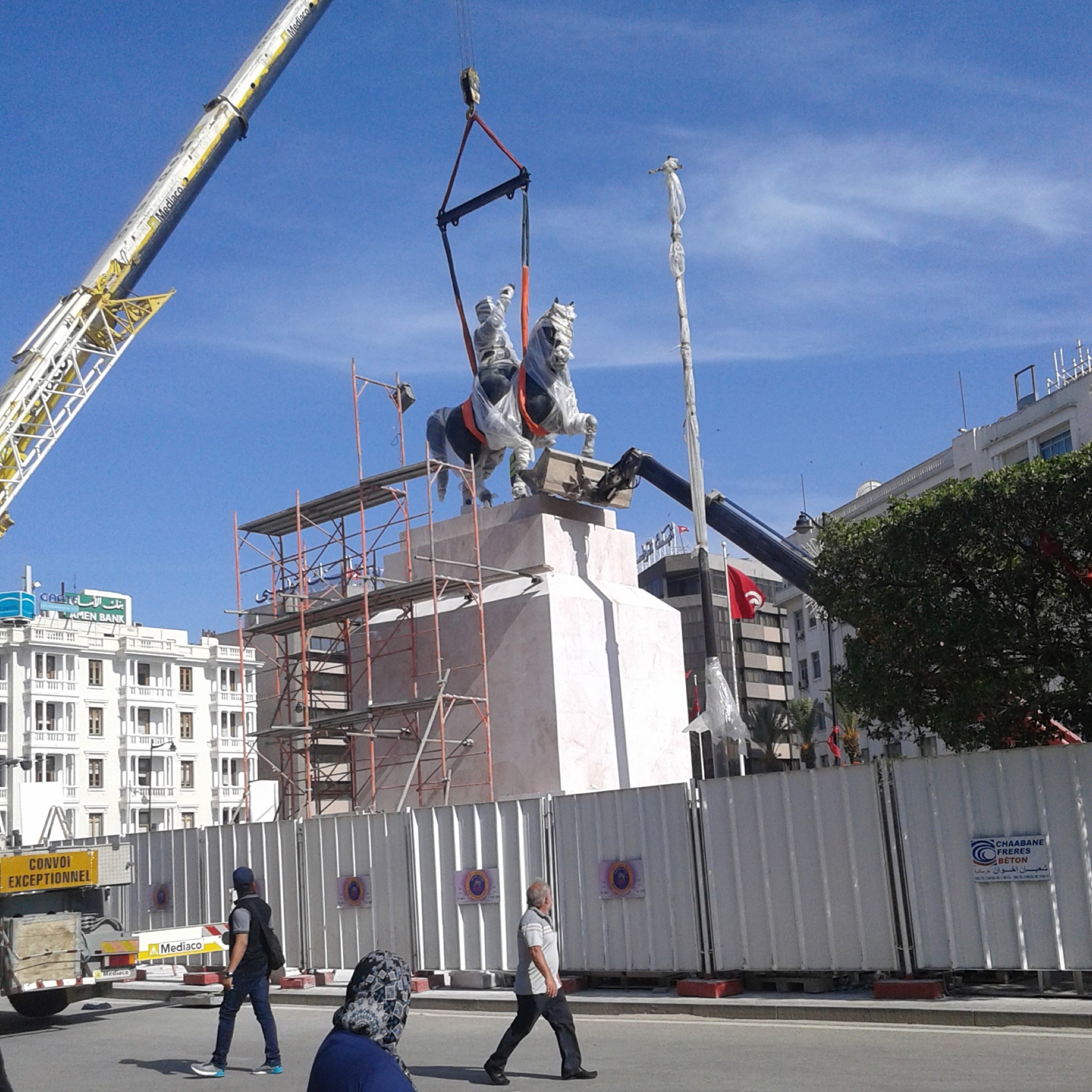 L'image montre un monument en cours d'installation. On peut voir une statue équestre accompagnée d'un cavalier, qui est en train d'être soulevée par une grue. Des échafaudages entourent la base de la statue, et un panneau de chantier est visible en arrière-plan. Le ciel est bleu, indiquant une journée ensoleillée. Des personnes marchent au premier plan, certaines portant des vêtements traditionnels.