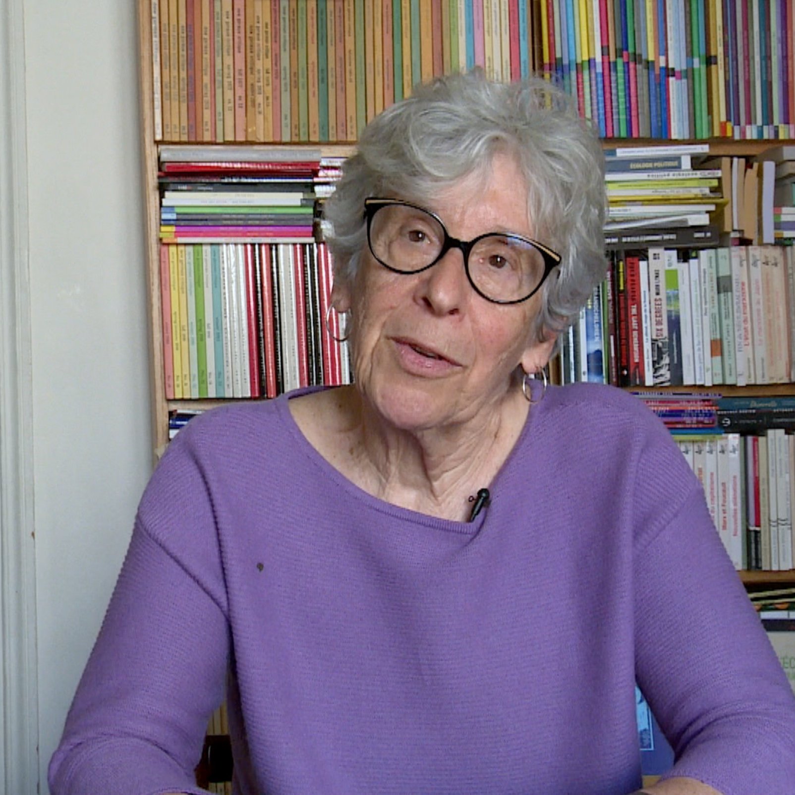 The image features an elderly woman with gray hair and glasses, sitting at a table. She is wearing a purple sweater and appears to be engaged in conversation or sharing her thoughts. In the background, there are bookshelves filled with various books, creating a colorful and organized backdrop. The setting suggests a cozy and intellectual atmosphere.