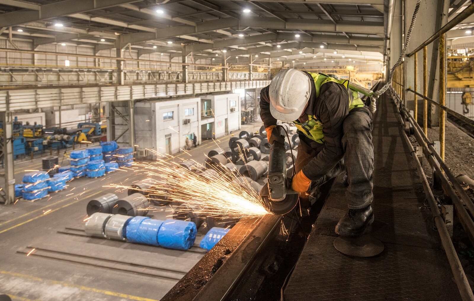 L'image montre un ouvrier en train de travailler dans un environnement industriel, probablement dans une usine. Il utilise une meuleuse, ce qui génère des étincelles. L'ouvrier porte un casque de sécurité et des gants, ce qui indique qu'il suit des mesures de sécurité adéquates. En arrière-plan, on peut voir des bobines de matériaux, ainsi que différents équipements et machines typiques d'un espace de travail industriel. L'éclairage de l'atelier semble naturel, avec des zones bien éclairées et d'autres plus sombres, mettant en valeur le côté dynamique et actif de l'environnement de travail.