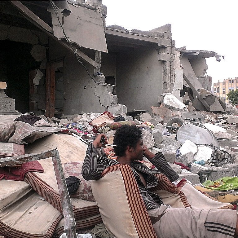 L'image montre une scène de destruction, probablement après un conflit ou une catastrophe naturelle. Au premier plan, un homme assis sur des matelas usés semble perdu dans ses pensées, entouré de débris. À l'arrière-plan, des bâtiments partiellement effondrés et des gravats jonchent le sol, témoignant des conséquences dévastatrices de l'événement. L'atmosphère est sombre et tragique, avec une sensation de désolation.