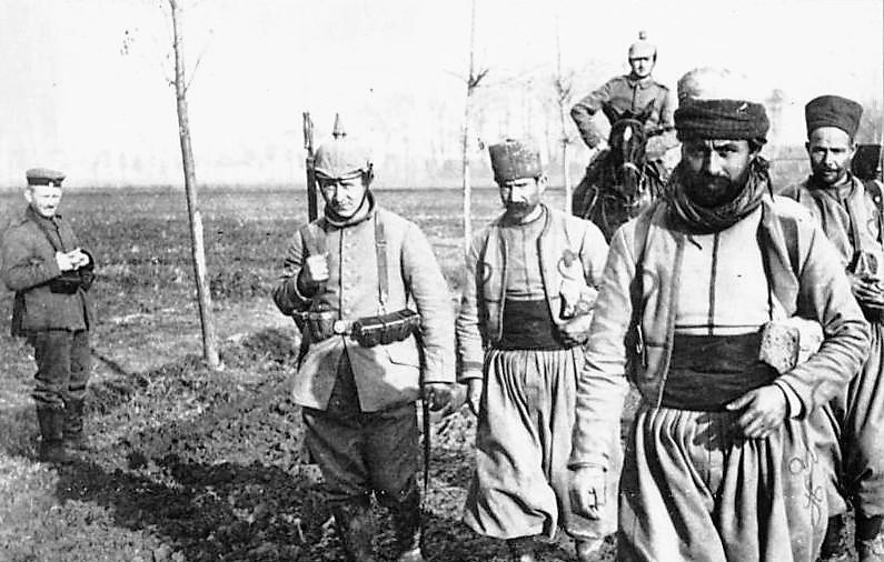 L'image montre un groupe de soldats marchant sur un terrain boueux. Ils portent des uniformes traditionnels, comprenant des pantalons larges et des bonnets. L'un des soldats est monté sur un cheval, tandis qu'un autre se tient en retrait, à l'écart du groupe. Le décor semble rural, avec quelques arbres en arrière-plan, suggérant un contexte militaire historique. L'atmosphère est sérieuse et solennelle, reflétant les conditions de la guerre.
