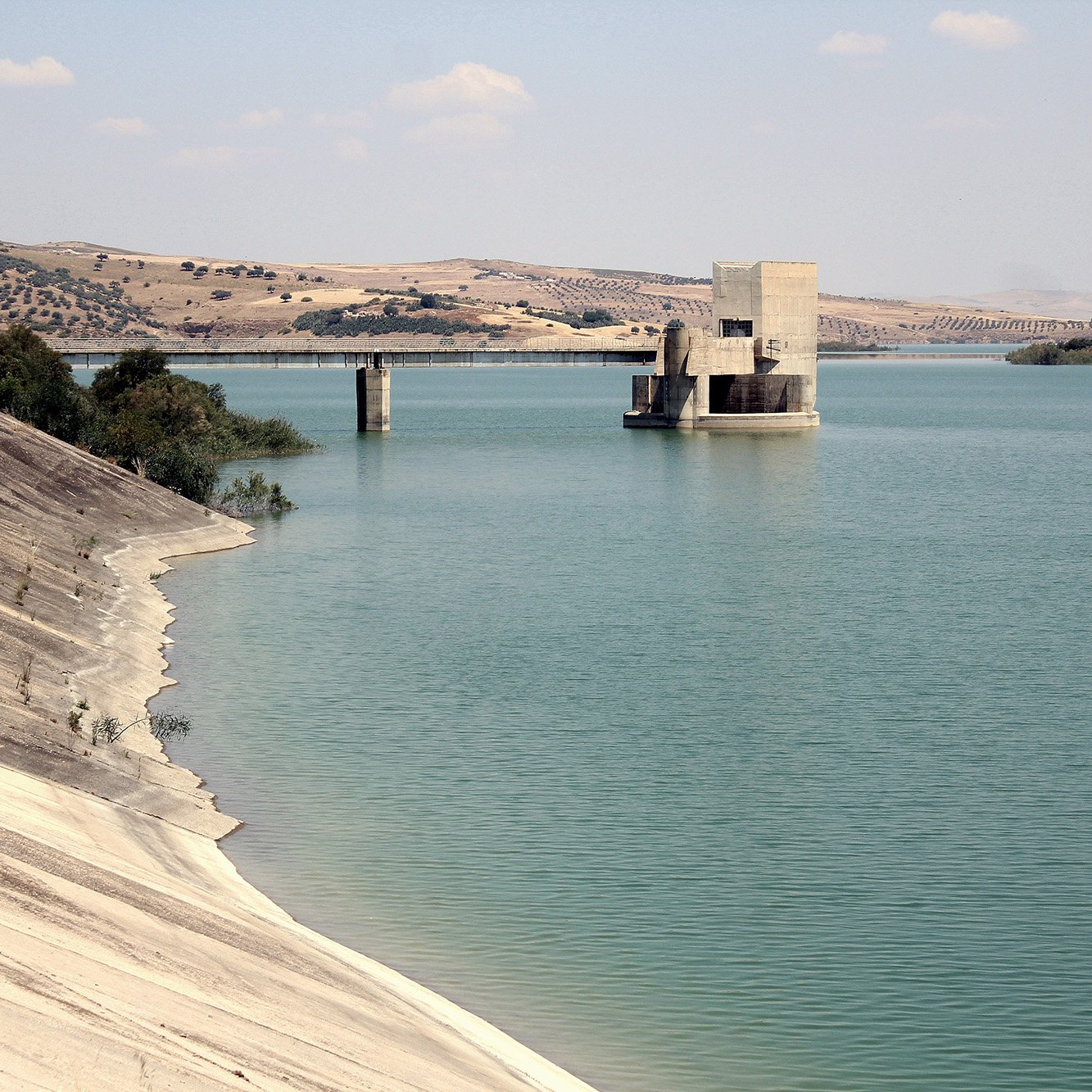 تظهر الصورة منظرًا هادئًا لبحيرة محاطة بتضاريس جبلية. يتوسط المشهد بناء هندسي يُحتمل أن يكون محطة لتحكم في المياه أو وسيلة لتخزينها. المياه باللون الأزرق الفاتح تعكس السماء، بينما تمتد ضفاف البحيرة بلطف، مع وجود تدرجات من اللون الرمادي الفاتح في الرمال. الأفق يكتمل بكتل من التلال الخضراء البعيدة، مما يضفي على الصورة جمالًا طبيعيًا وهدوءً.