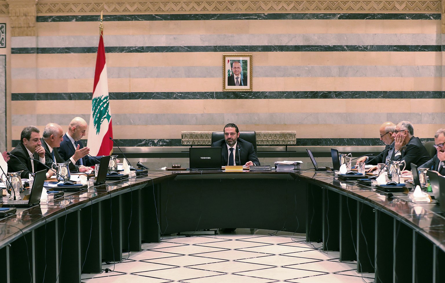 L'image montre une réunion dans une salle de conférence. Au centre, une grande table ovale est entourée de plusieurs personnes vêtues de costumes et cravates. À l'arrière-plan, on peut voir un drapeau libanais et un portrait, probablement du président du Liban, accroché au mur. Les participants semblent être à l'écoute et engagés dans la discussion, avec des documents et des ordinateurs portables devant eux. L'ambiance semble formelle et professionnelle.