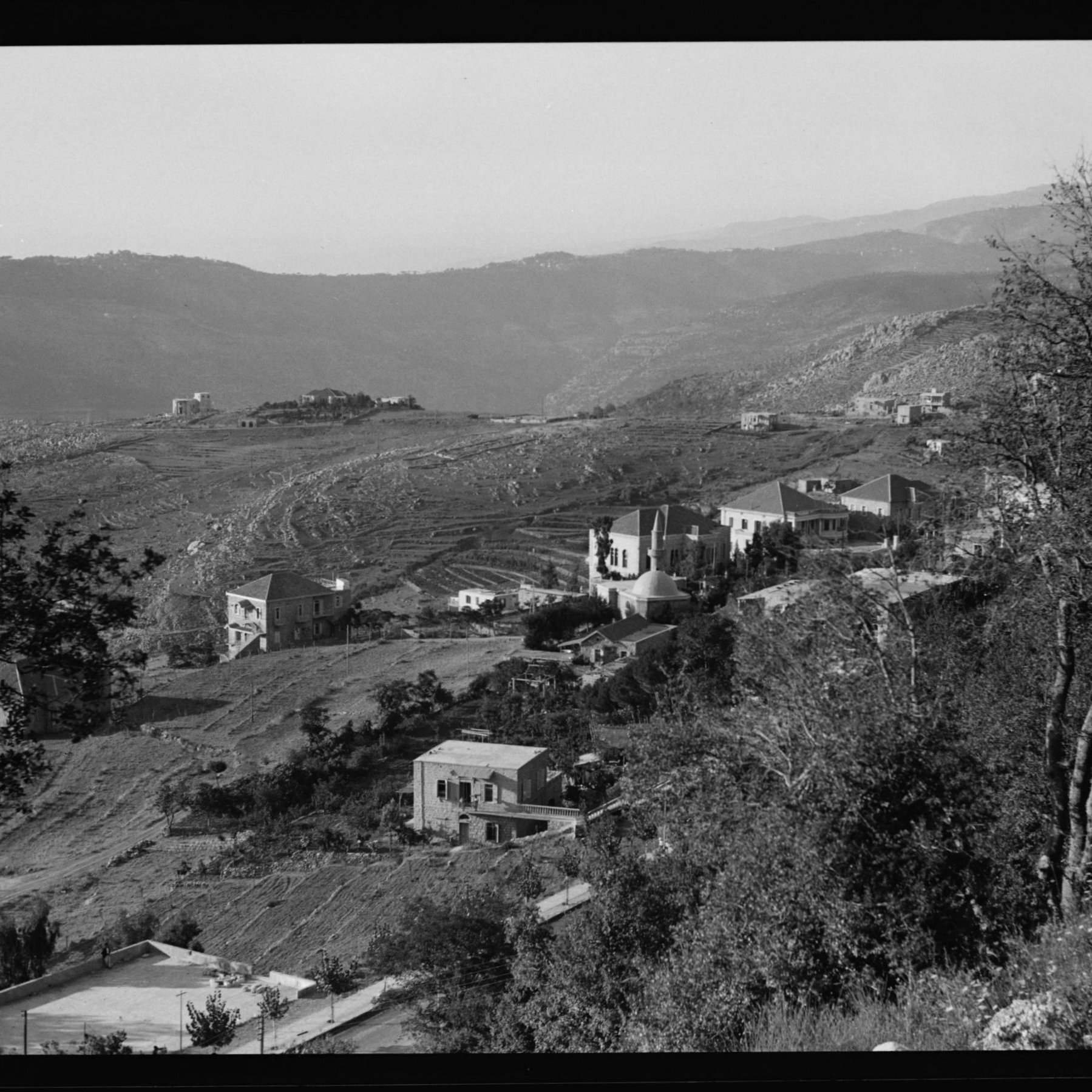 L'image montre un paysage montagneux avec des collines qui s'étendent à l'horizon. On peut apercevoir plusieurs maisons éparpillées sur les flancs des collines, entourées de végétation. La scène semble être prise en noir et blanc, ce qui lui donne un aspect nostalgique. Les terrains agricoles sont visibles, indiquant une zone rurale. Les montagnes en arrière-plan ajoutent une profondeur à la composition paysagère.