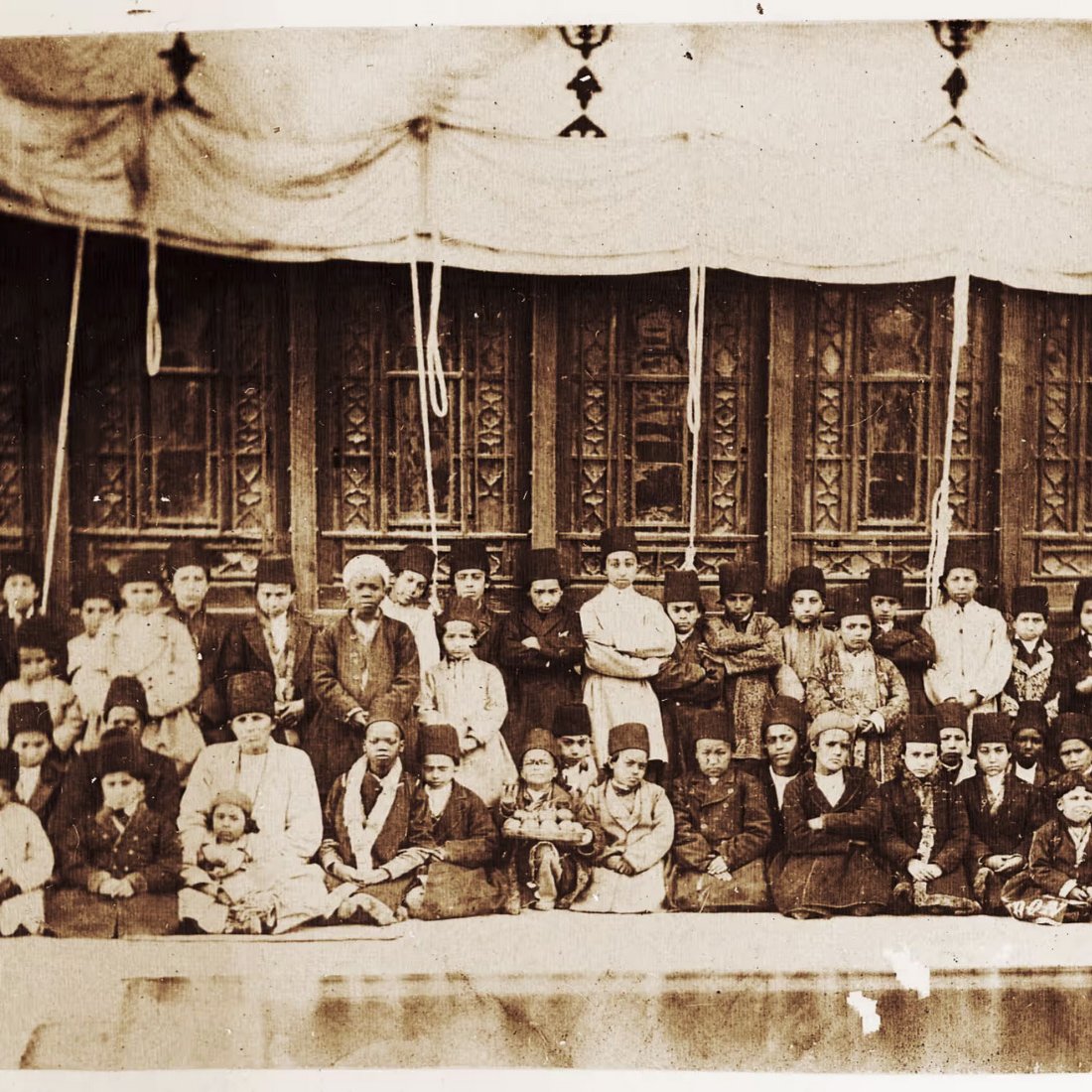 L'image présente un groupe de personnes, probablement une photographie en noir et blanc, où l'on peut voir une grande assemblée d'hommes, de femmes et d'enfants. Ils sont assis ou debout devant un bâtiment avec des fenêtres ornées et des décorations. Les vêtements portés semblent typiques d'une époque passée, suggérant un contexte historique. L'ensemble donne une impression de communauté ou d'événement social.