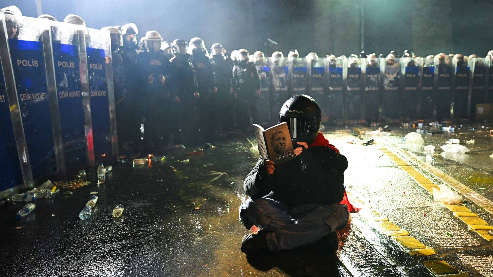 Un manifestant, masqué, lit un livre, face à des policiers en tenue anti-émeute.
