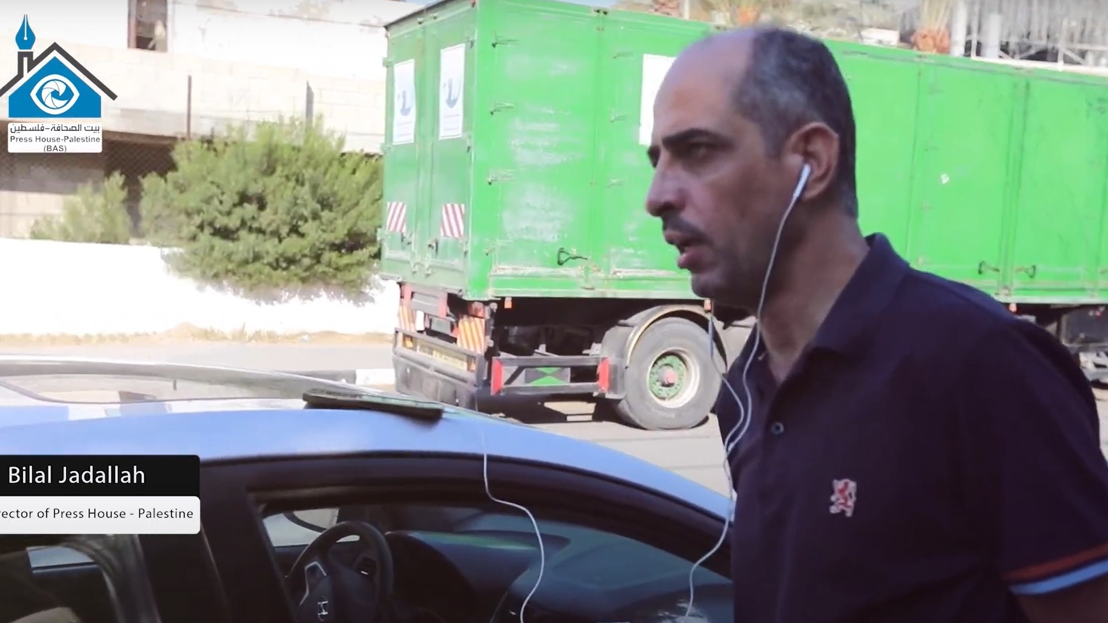 Nell'immagine si vede un uomo che cammina accanto a un'auto. Indossa una maglietta scura e ha delle cuffie che gli pendono dalle orecchie. Sullo sfondo si nota un camion verde e alcune piante. L'uomo sembra concentrato e diretto verso un punto specifico. A sinistra, è presente un'etichetta che indica il suo nome e la sua posizione, come direttore di un'organizzazione legata alla stampa in Palestina.