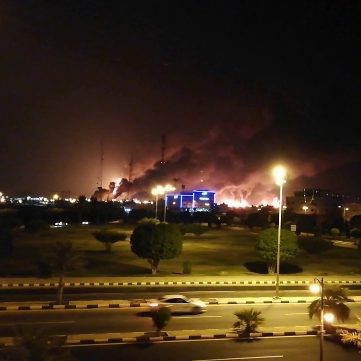 L'image montre une scène nocturne où l'on peut voir des nuages de fumée s'élevant dans le ciel, suggérant qu'il y a un incendie ou une explosion quelque part à l'horizon. Les lumières des bâtiments environnants sont visibles, créant un contraste avec l'obscurité de la nuit. La route devant est éclairée, avec quelques voitures qui passent. Des arbres et des espaces verts se trouvent également au premier plan, ajoutant une certaine profondeur à la scène.