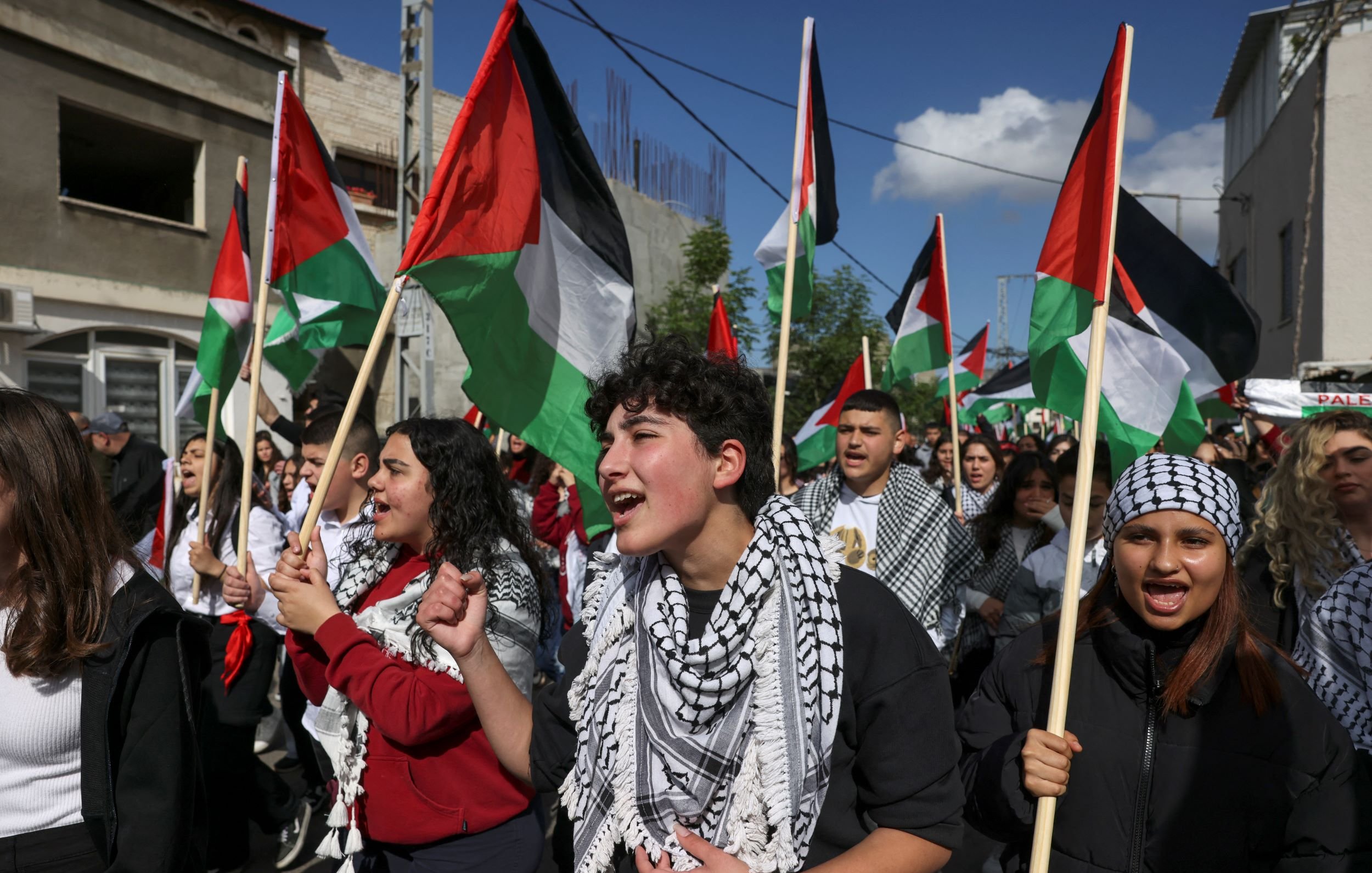 L'immagine mostra un gruppo di persone che partecipano a una manifestazione. Molti manifestanti tengono in mano bandiere palestinesi, caratterizzate da strisce nere, bianche e verdi e da un triangolo rosso. Alcuni di loro indossano kefiah, tradizionali sciarpe palestinesi. Le espressioni sui loro volti trasmettono passione e determinazione mentre cantano o gridano slogan, contribuendo a creare un'atmosfera di unità e attivismo. Sullo sfondo, si intravedono edifici e il cielo è parzialmente nuvoloso.