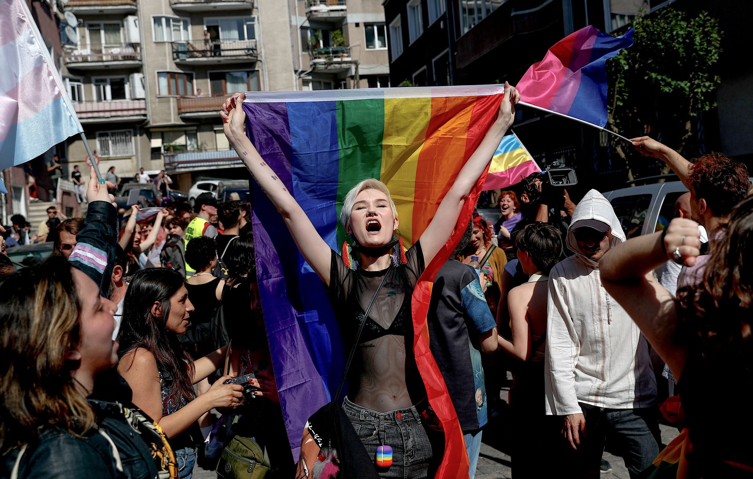 L'immagine mostra un gruppo di persone in una strada affollata, probabilmente durante una manifestazione o una parata. Al centro, una giovane donna con capelli corti e bianchi è in primo piano, mentre tiene alta una grande bandiera arcobaleno. Sembra esprimere gioia e determinazione. Intorno a lei, altre persone sventolano bandiere e manifestano il loro sostegno, creando un'atmosfera di festeggiamento e orgoglio. Le decorazioni e i colori vivaci trasmettono un messaggio di inclusione e solidarietà.
