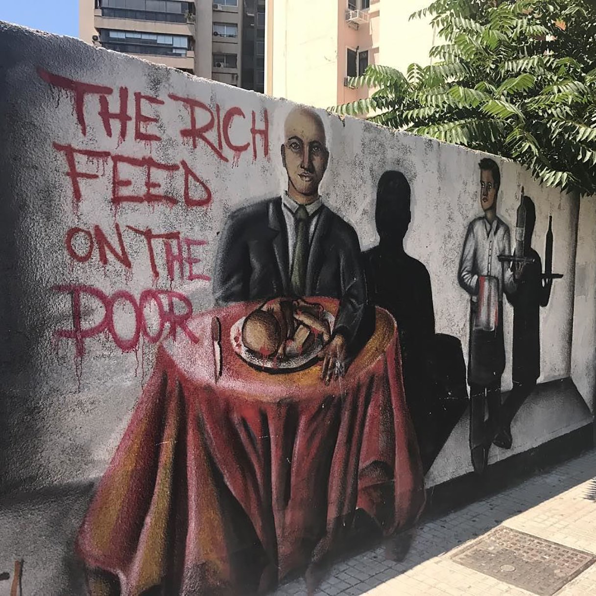 L'image montre une fresque murale colorée sur un mur. Au premier plan, un homme en costume est assis à une table couverte d'une nappe rouge, avec un plat à manger devant lui. À côté, on peut voir une ombre d'un serveur, suggérant un contexte de service alimentaire. Sur le mur, il est écrit "THE RICH FEED ON THE POOR", ce qui évoque un message critique sur les inégalités sociales. Des éléments de l'arrière-plan, comme des bâtiments et de la verdure, ajoutent à l'environnement urbain.