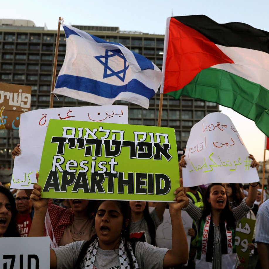 تظهر الصورة تجمعًا حاشدًا لمتظاهرين يحملون شعارات ولافتات. في الخلفية، هناك علم إسرائيلي وعلم فلسطيني. يرفع المتظاهرون شعارات تدعو لمقاومة الفصل العنصري. الأجواء تعكس روح الاحتجاج والمطالبة بالحقوق.
