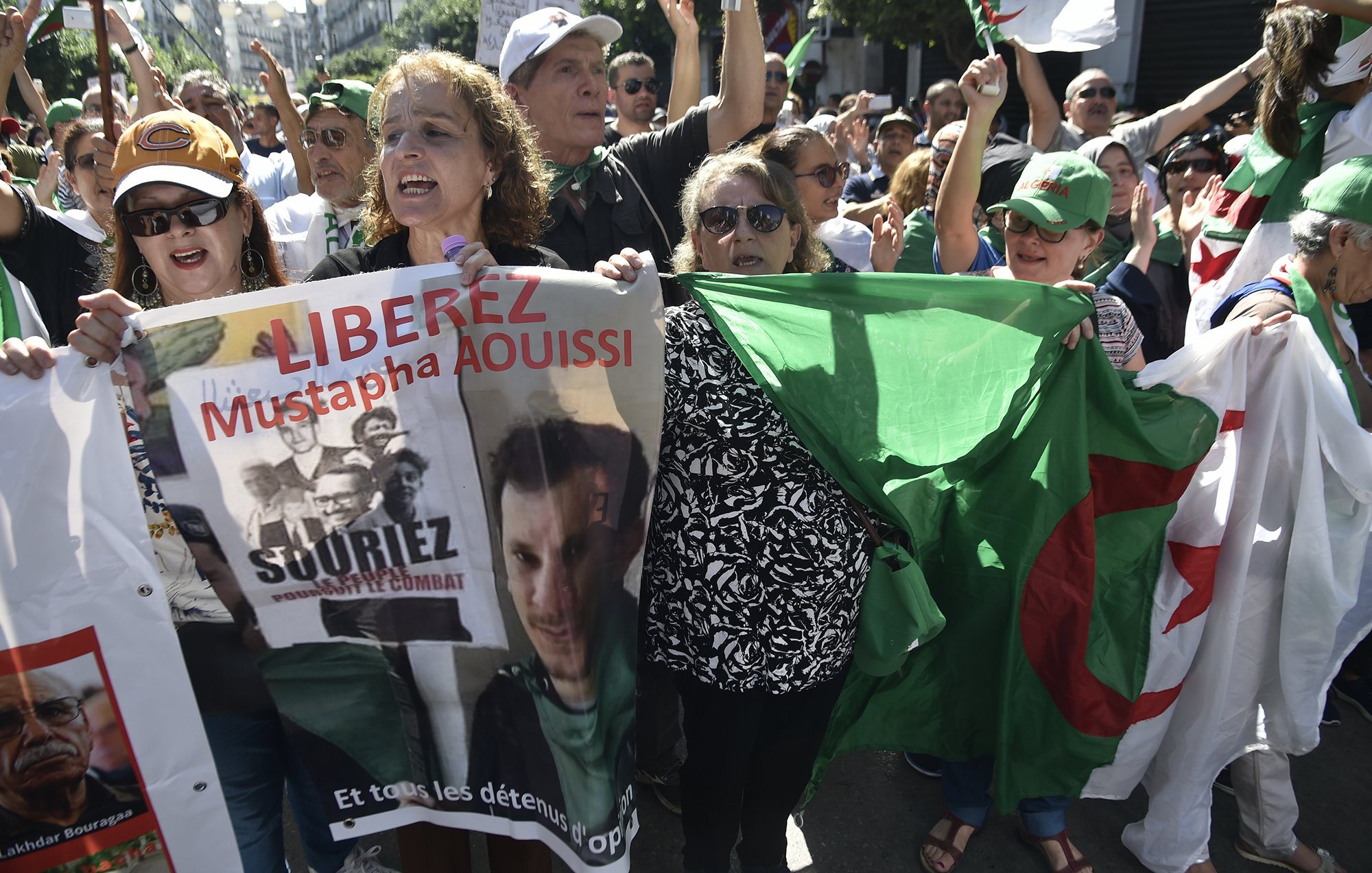 L'image montre une manifestation où des personnes sont rassemblées, exprimant des revendications. On peut voir des individus brandissant des pancartes et des drapeaux, notamment un drapeau algérien. Les manifestants semblent mobilisés et engagés, scandant des slogans pour la libération de Mustapha Aouissi. L'atmosphère semble dynamique et pleine d'énergie, avec une forte présence de diversité parmi les participants.