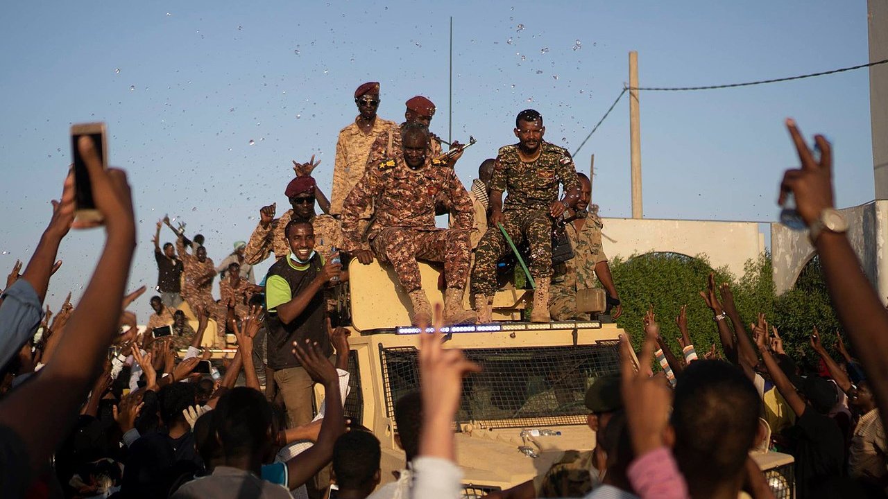 الصورة تظهر مجموعة من الجنود يقفون على متن مركبة عسكرية، بينما يبدو أن حشودًا من الناس تتجمع حولهم. يبدو الجو احتفاليًا حيث يرفع بعض الأشخاص أيديهم في الهواء، ويبدو أنهم يعبرون عن الفرح أو التأييد. السماء زرقاء وظهرت بعض الأعلام والاحتفالات في الخلفية.