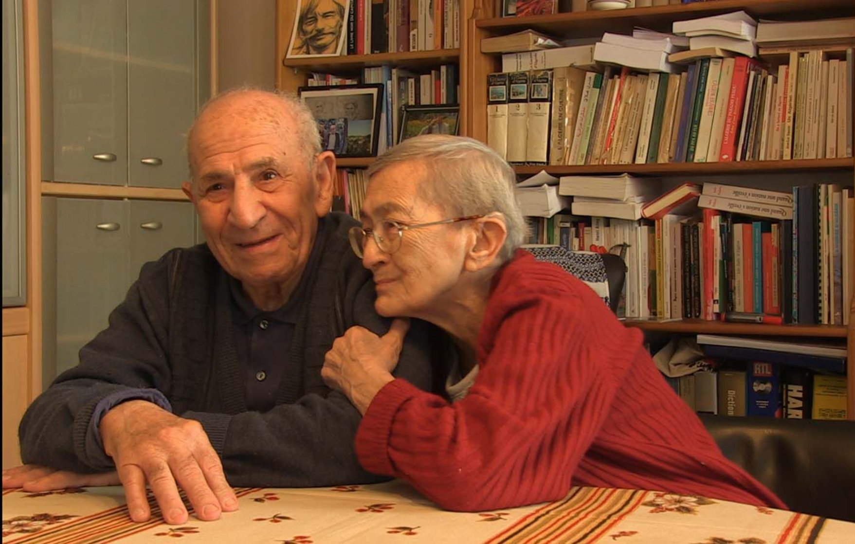 L'image montre un couple âgé assis à une table. L'homme a les cheveux gris et porte un pull sombre, souriant avec bienveillance. La femme, également âgée, a des cheveux très courts et porte un pull rouge. Elle est légèrement penchée vers lui, affichant une expression tendre et affectueuse. En arrière-plan, on peut apercevoir des étagères remplies de livres, créant une atmosphère chaleureuse et intellectuelle. La décoration présente un style simple et accueillant.