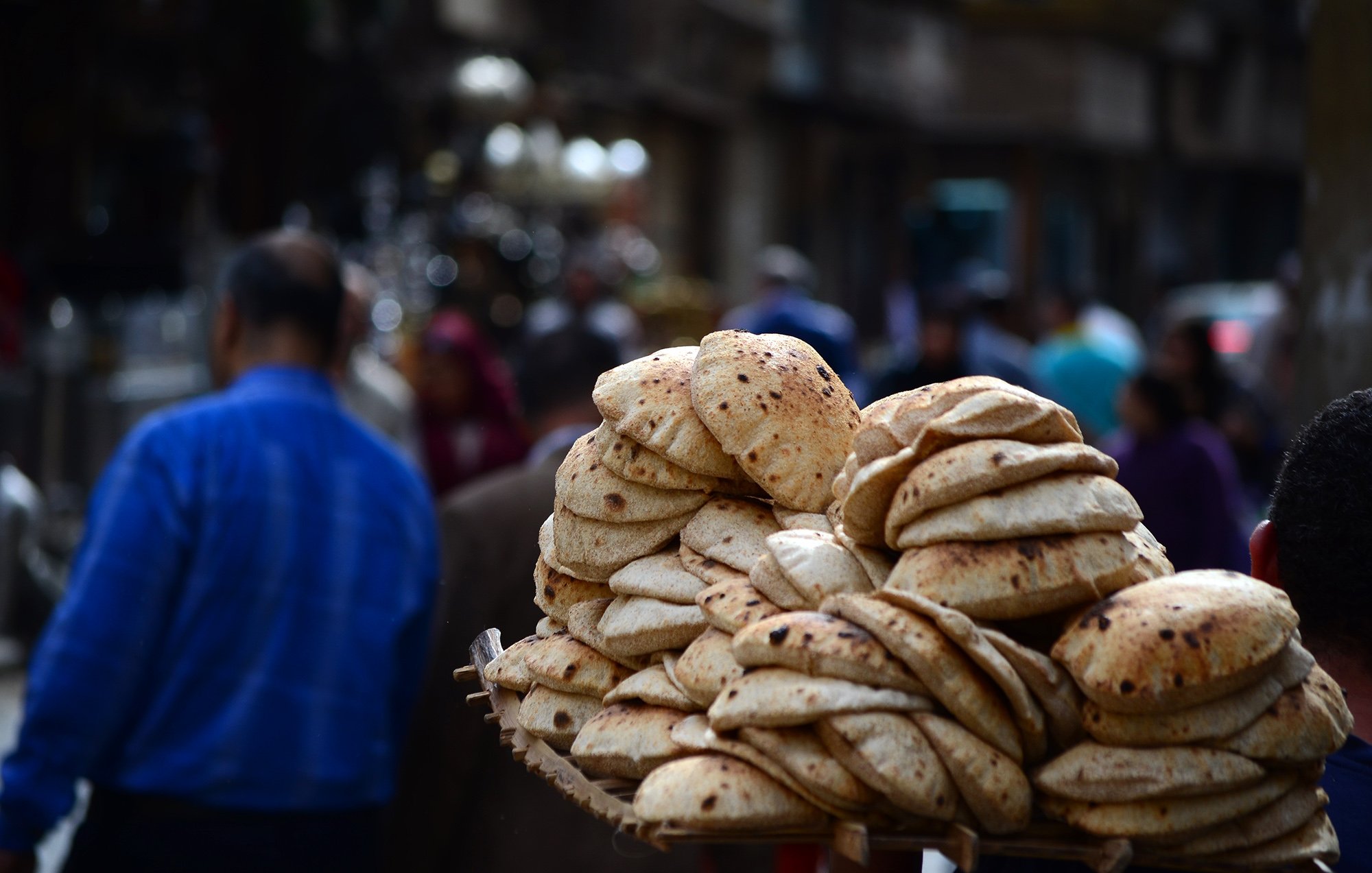 تظهر الصورة مجموعة من الخبز الطازج، يبدو أنه خبز عربي، مُتراص بشكل جميل في سلة يحملها شخص. في الخلفية، يتواجد عدد من الأشخاص في شارع مزدحم، مما يوحي بأجواء نابضة بالحياة في المدينة. يركز المشهد على الخبز كجزء من الثقافة الغذائية.