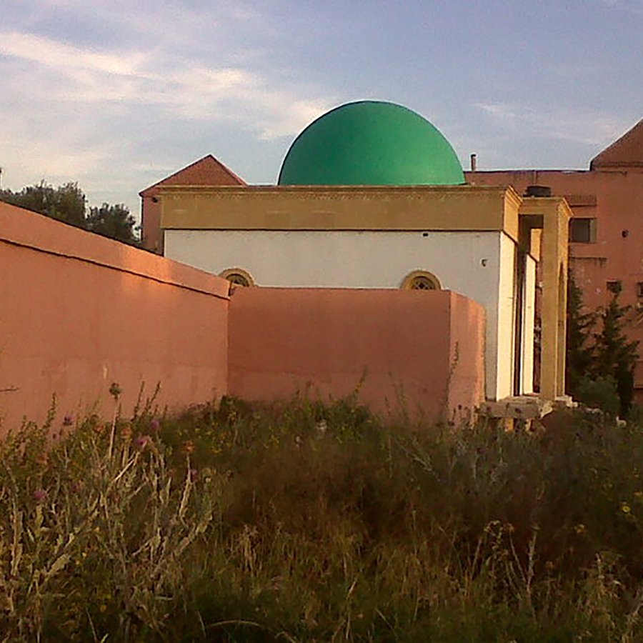L'image montre un bâtiment avec une coupole verte, partiellement cachée par un mur. Le bâtiment semble être de style architectural traditionnel, avec des murs peints en rose et des fenêtres rondes. On remarque également une végétation sauvage devant, ajoutant un aspect naturel à la scène. En arrière-plan, on aperçoit d'autres structures, suggérant un environnement urbain ou semi-urbain. Le ciel est clair, indiquant une atmosphère paisible.