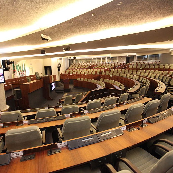 L'image montre une salle de réunion ou un hémicycle, probablement utilisée pour des sessions parlementaires ou des assemblées. On peut voir des rangées de sièges confortables disposés en arc de cercle, avec des tables en bois devant chaque siège. Des écrans et des équipements audiovisuels sont visibles, indiquant que la salle est équipée pour des présentations ou des débats. Les murs sont décorés de manière sobre et moderne, créant une ambiance institutionnelle.