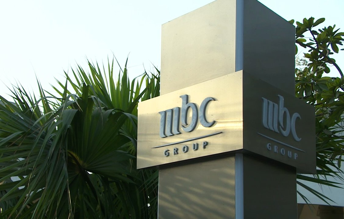 L'image montre un panneau en métal portant le logo de "MBC Group". Le panneau est installé dans un environnement extérieur, entouré de végétation, notamment des palmiers. Le fond est clair, suggérant une ambiance ensoleillée.