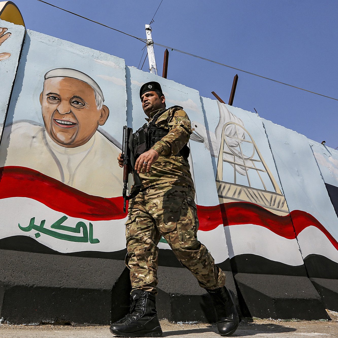 L'image montre un soldat en uniforme, armé et en marche, devant un mur peint. Sur le mur, il y a une fresque représentant un homme souriant et levant la main, probablement symbolisant une figure importante ou un leader. Le fond de la fresque inclut des éléments décoratifs et des couleurs qui évoquent le drapeau irakien. L'environnement semble être une zone urbaine, marquée par des installations de sécurité.