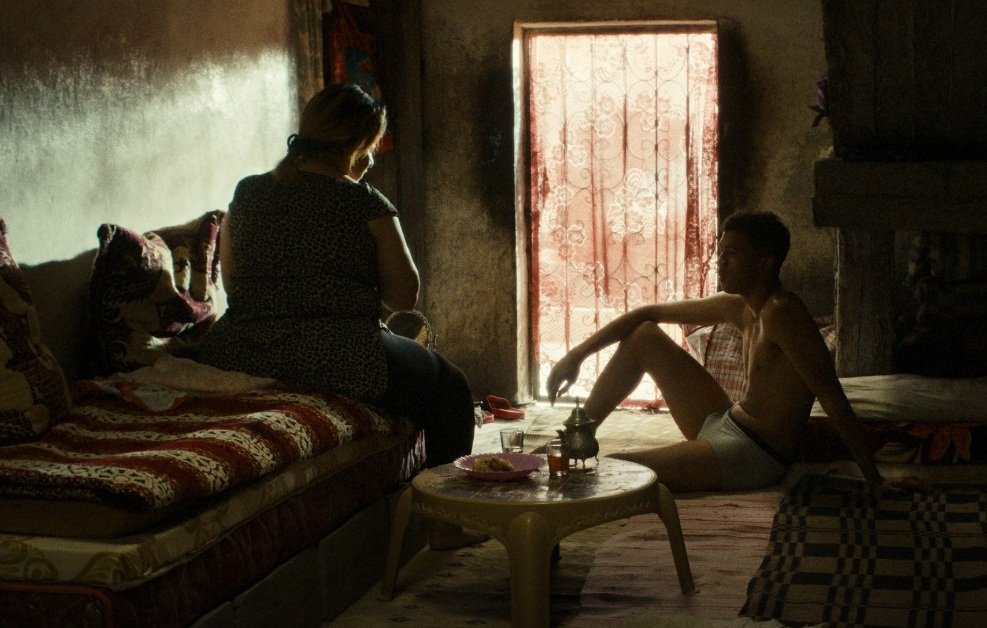 L'image montre une scène intérieure, probablement dans un espace de vie modeste. On y voit deux personnes, assises dans une pièce faiblement éclairée par la lumière qui entre à travers une fenêtre avec des rideaux. Les murs sont de couleur sombre et le mobilier semble simple. L'une des personnes est assise sur un canapé, tandis que l'autre est assise par terre, semblant contempler la pièce ou se relaxer. L'atmosphère dégage une sensation d'intimité et de calme. Des objets sont présents, suggérant une vie quotidienne.