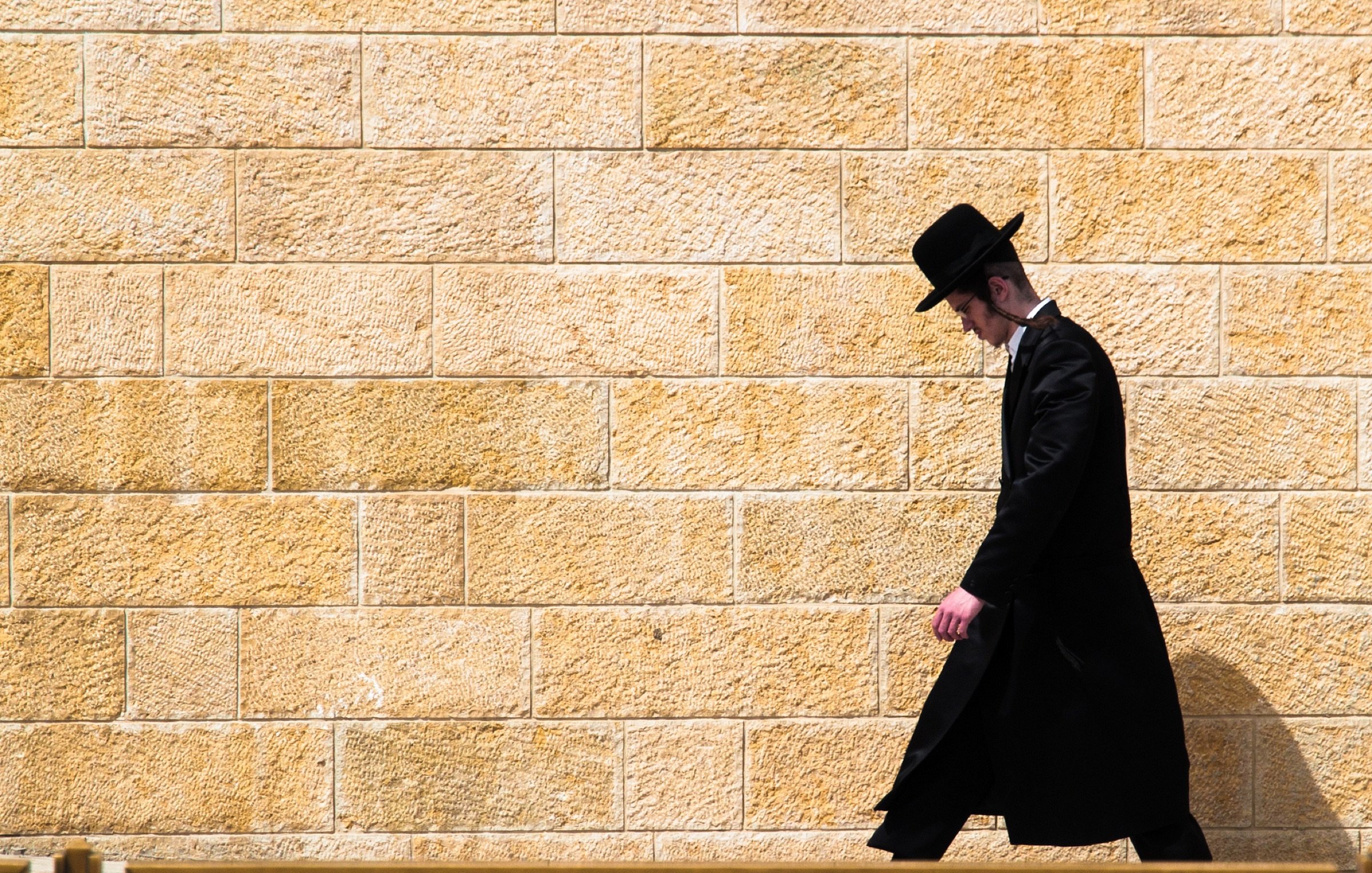 L'image montre un homme marchant le long d'un mur de pierre de couleur beige. Il est habillé d'un long manteau noir et porte un chapeau noir. Son expression est sérieuse et il semble concentré sur son chemin. L'arrière-plan est uni, ce qui met en valeur la silhouette de l'homme qui avance seul.