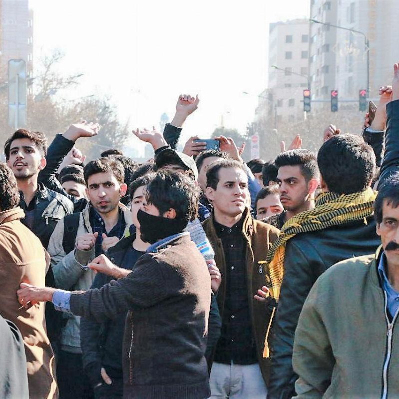 L'image montre une foule de personnes rassemblées dans une rue. Les individus semblent engagés dans une forme de manifestation ou de rassemblement collectif. Certains lèvent les mains ou filment avec des téléphones, tandis que d'autres ont des expressions préoccupées ou déterminées. On peut voir une diversité d'habillements, y compris des vêtements traditionnels, et un mélange de genres parmi les participants. L'ambiance générale semble être celle de l'enthousiasme ou de la passion pour une cause commune.