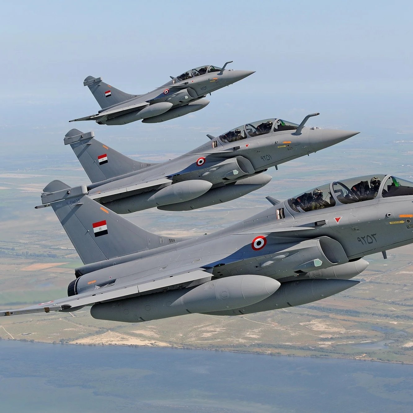 L'image montre un groupe de jets de chasse en plein vol. Trois avions de chasse, probablement des Rafales, sont alignés en formation, avec des couleurs principalement grises. Chaque avion porte des marqueurs et des emblèmes, semblant appartenir à une force aérienne. Le décor en arrière-plan indique un paysage vaste, avec des zones de terre et de l'eau visible, suggérant un vol à basse altitude. L'ensemble dégage une impression de puissance et de précision militaire.