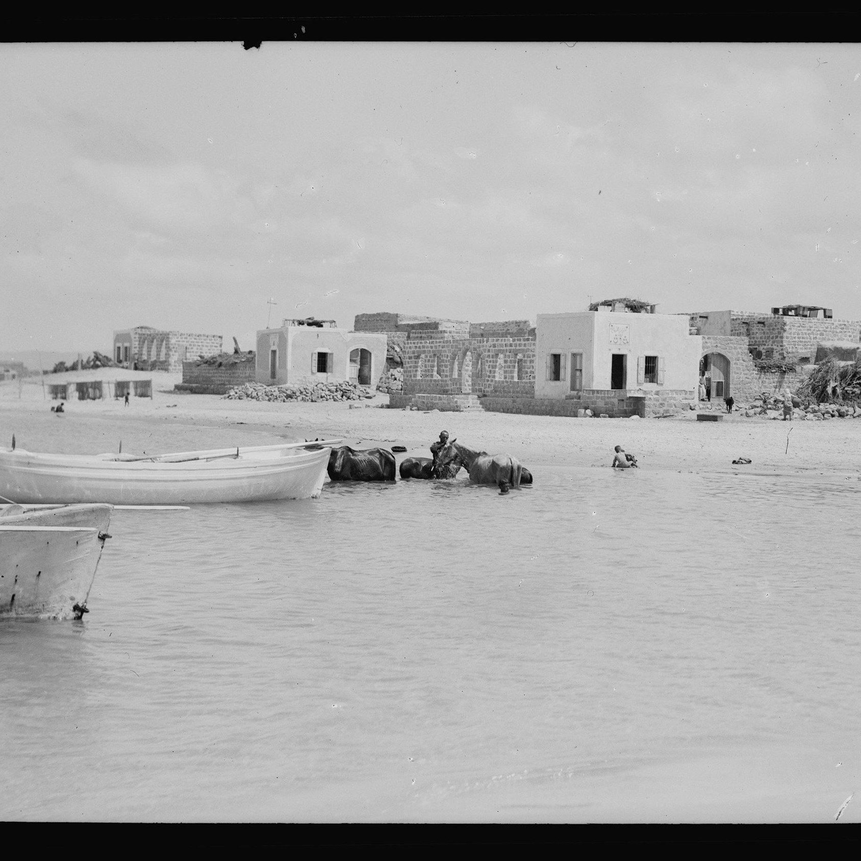 تظهر في الصورة مشهداً ساحلياً يضم مجموعة من المراكب الصغيرة الراسية على المياه. في الخلفية، توجد مبانٍ قديمة إلى جانب بعض الهياكل المتبقية، ما يدل على وجود مجتمع ساحلي سابق. يُرى بعض الأشخاص، بما في ذلك شخص يركب على ظهر حيوان، مما يعطي انطباعًا عن الحياة اليومية في هذا المكان. تتسم السماء بالغيوم المتفرقة، مما يوحي بأن الصورة ربما تم التقاطها في يوم مشمس.