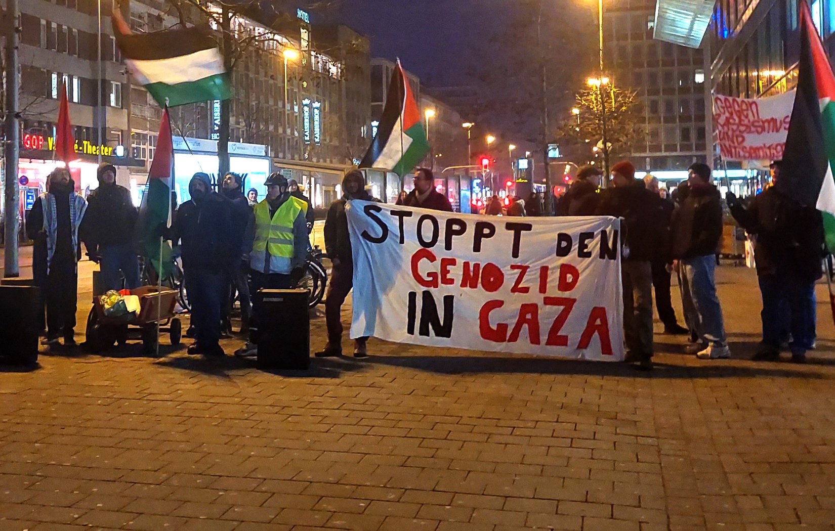L'image montre un groupe de manifestants rassemblés dans une rue, tenant une grande banderole sur laquelle il est écrit "STOPPT DEN GENOZID IN GAZA". Ils portent des drapeaux palestiniens et semblent exprimer leur soutien à Gaza. L'atmosphère est sérieuse, et il est probable que la manifestation s'inscrive dans un contexte de protestation contre des violences ou des injustices en Palestine. Les lumières des bâtiments environnants suggèrent qu'il s'agit d'une manifestation en soirée.