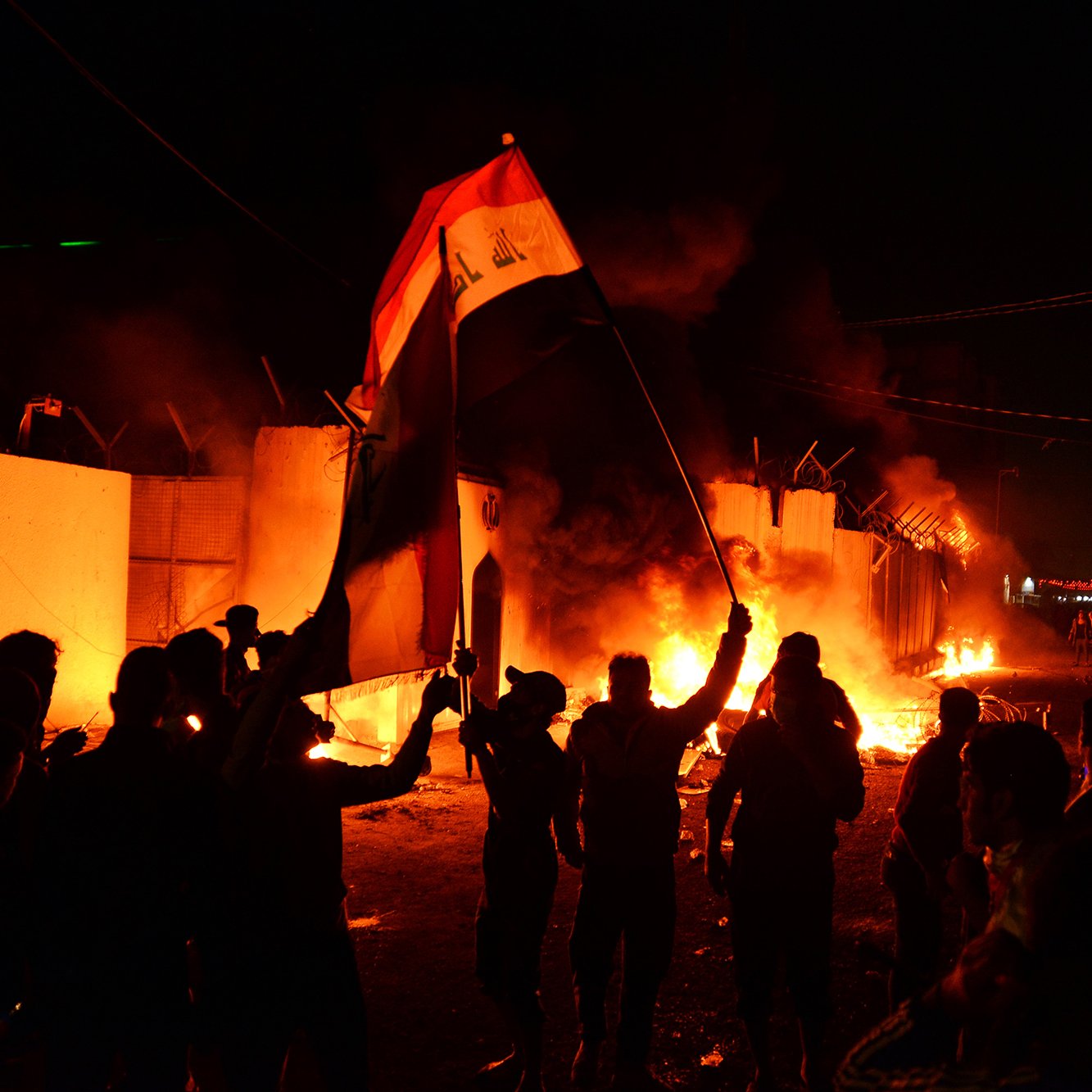 L'image montre une scène nocturne animée, probablement lors d'une manifestation ou d'une émeute. Des personnes brandissent un drapeau, en l'occurrence le drapeau irakien, tandis que des flammes s'élevées en arrière-plan, suggérant des tensions ou des troubles. Les silhouettes des manifestants sont bien visibles, et l'ambiance est chargée d'énergie et d'émotion. La lumière des feux crée un contraste fort avec l'obscurité de la nuit.