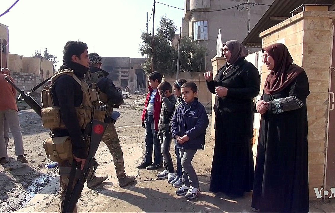 تظهر في الصورة مجموعة من الأشخاص في شارع يبدو أنه في منطقة تتعرض للتوتر أو النزاع. في الجهة اليسرى، يوجد جندي مسلح يرتدي زيًا عسكريًا، ويتحدث مع مجموعة من النساء والأطفال. النساء يرتدين ملابس تقليدية، بينما يبدو الأطفال مهتمين بالمحادثة. خلفهم، يمكن رؤية مباني متضررة وأجواء تعكس حالة المنطقة.