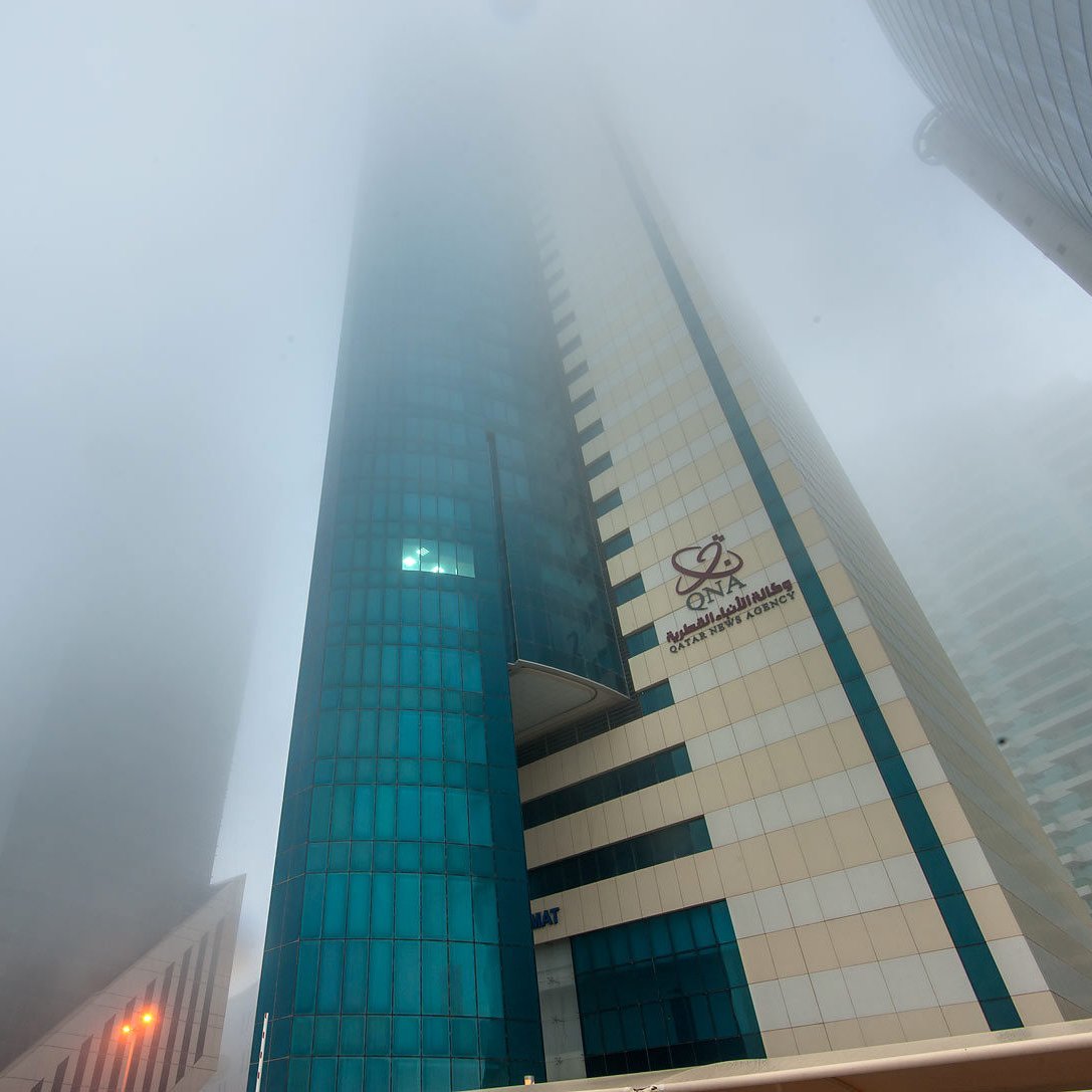 L'image montre un paysage urbain avec des gratte-ciels partiellement cachés par un épais brouillard. La visibilité est réduite, ce qui donne une atmosphère mystérieuse et peu lumineuse. Les bâtiments ont des façades modernes, avec des reflets de verre. On peut distinguer des éléments architecturaux, mais l'intensité du brouillard rend difficile l'observation claire des détails. Les lumières des rues sont visibles, créant un contraste avec l'environnement brumeux.