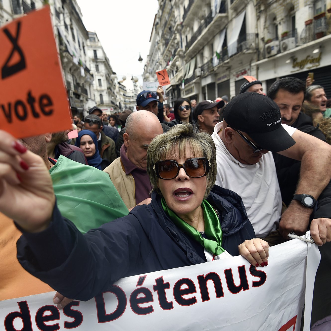 The image depicts a crowd of protesters gathered in an urban setting, likely during a demonstration. A woman in the foreground is holding an orange sign that reads "l'vote" while wearing sunglasses and a green top. The crowd behind her appears to be a mix of men and women, some holding signs and banners. The atmosphere conveys a sense of urgency and solidarity among the protesters, who seem to be advocating for a political cause. The background includes urban architecture, suggesting a city environment.