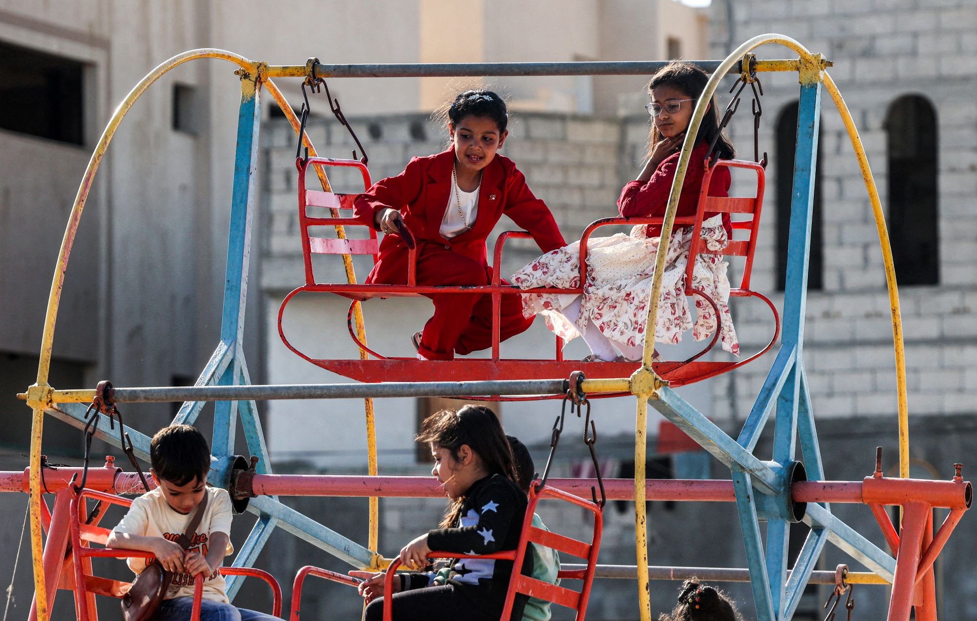 L'immagine mostra un'area giochi all'aperto, con una giostra colorata. Alcuni bambini sono seduti su seggiolini rossi mentre si divertono. Uno dei bambini indossa un completo rosso e sembra divertirsi molto, mentre un'altra bambina, vestita con un abito bianco con fiori, osserva da un'altezza superiore. Gli altri bambini sono anche loro intenti a giocare, creando un'atmosfera di gioia e spensieratezza in un contesto urbano.